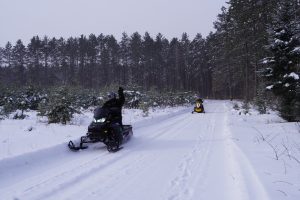 Two snowmobilers in the woods and the front person is waving at the camera