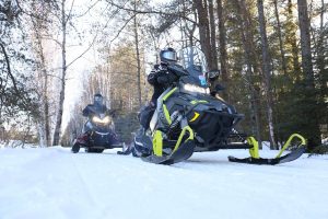 Looking up at two snowmobilers on a snowy path in the woods