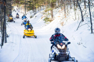 A group of people snowmobiling downhill in a snowy wooded area